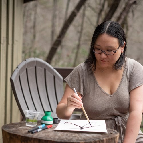 An Asian woman with shoulder length black hair and glasses in a gray-gold dress poses over a Korean brush painting on the deck of a cabin with a tree stump table. There is a forest in the background.